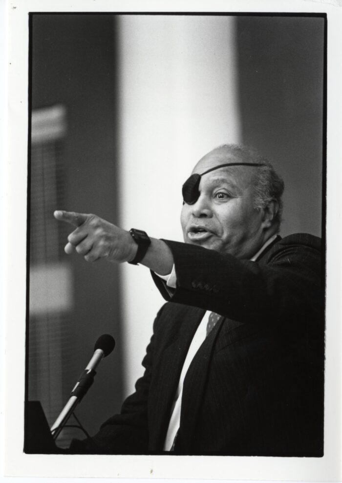 James Farmer, an older Black man wearing an eye patch, standing at a podium with a microphone. He is speaking and pointing outwards.