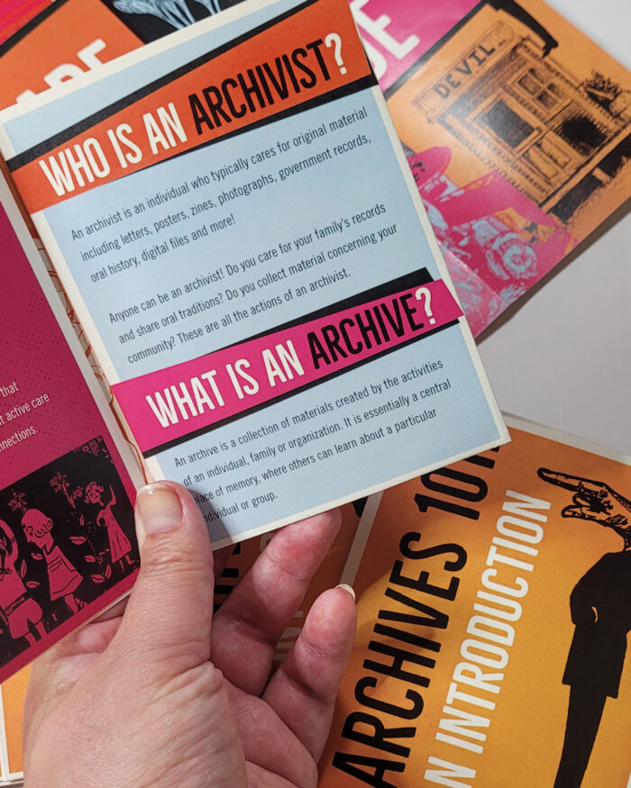 A person's hand holds open a zine to a page where the bold headers "Who is an archivist?" and "What is an archive" are prominent.