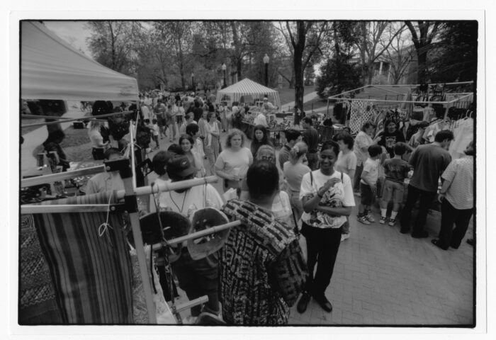 A large crowd of people gather at an outdoor fair. There are several tents and tables with vendors selling clothing and other items.