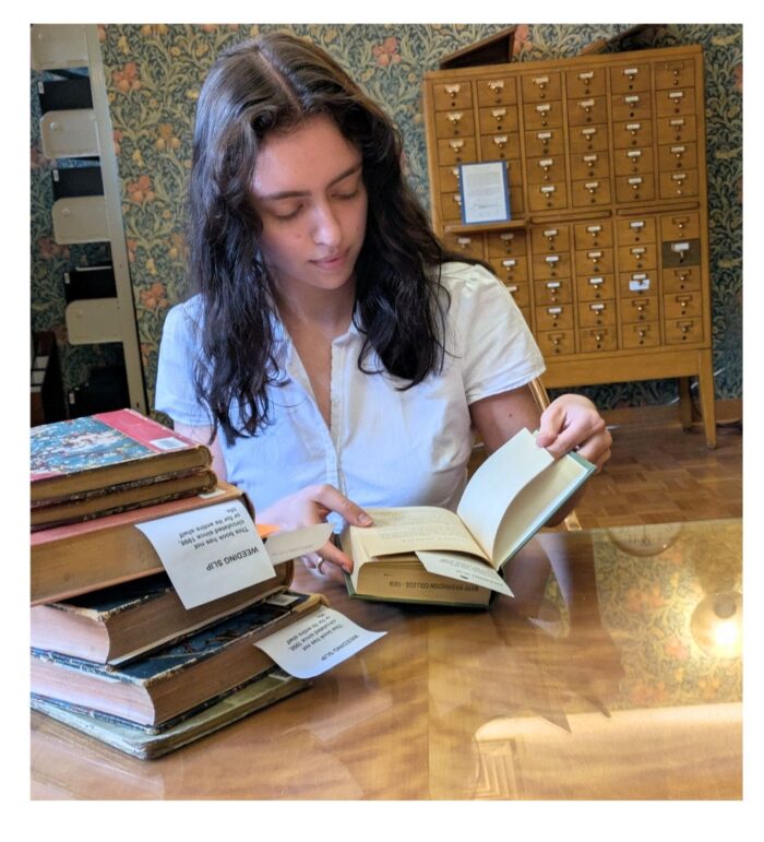 Person reading at a wooden table in a library, with stacked books, a card catalog, and shelves in the background.