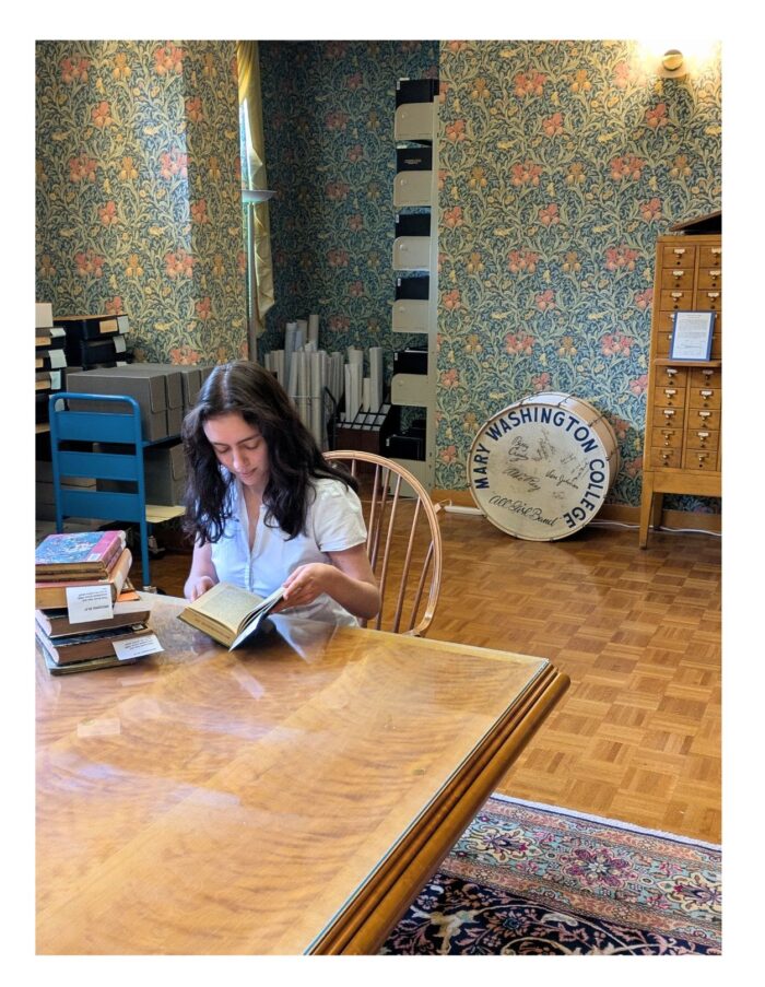 Person reading at a wooden table with books, surrounded by shelves of documents and a drum labeled ‘Mary Washington College.