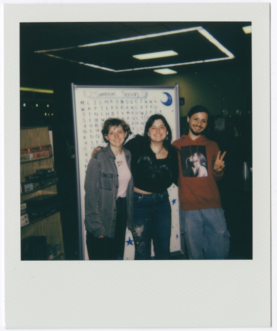 Polaroid of three people standing in front of a whiteboard; the person on the right makes a peace sign, with shelves visible in the background.