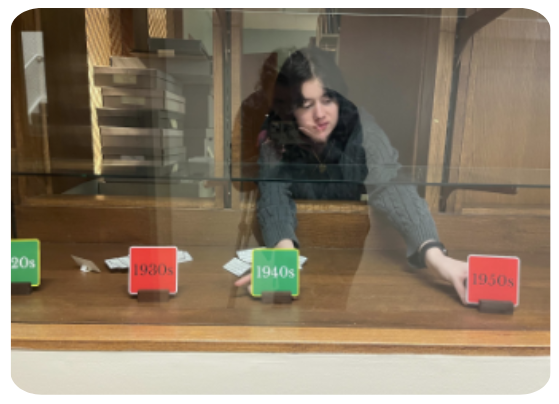 A student leans into a glass case while adjusting paper labels held up by wooden blocks. The paper labels read, from left to right, “20s, 1930s, 1940s, 1950s.”
