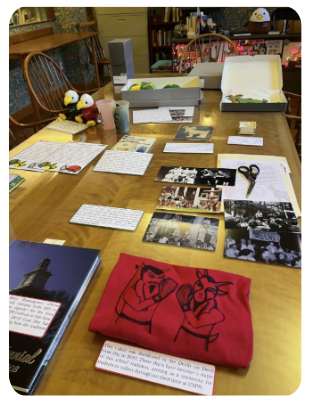 Artifacts and their labels arranged on a table. Red t-shirt, a yearbook, photographs, and eagle plushies wearing red and green colored shirts.