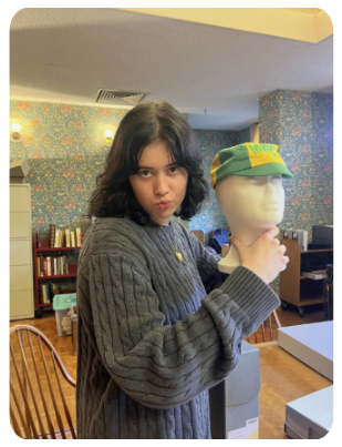Student in a blue sweater makes a puckered lips face while holding a mannequin head that is wearing a green and yellow cap with green letters that read “MWC.”