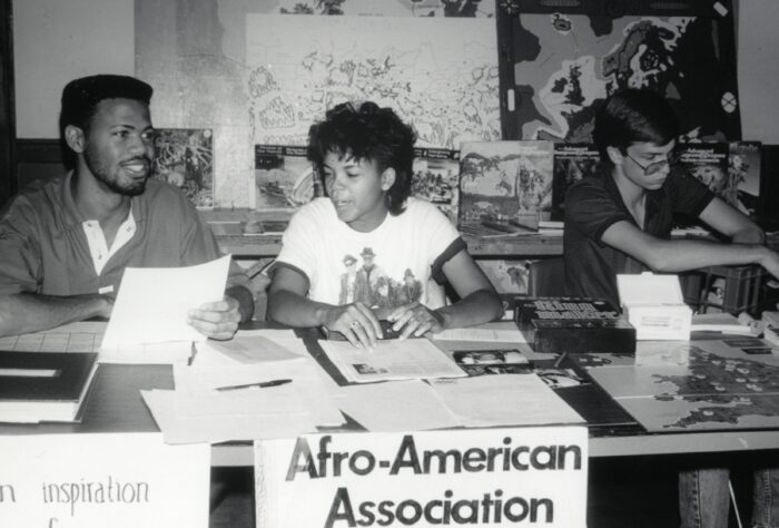 Three people sit at a table covered with papers, books, and informational materials. A sign on the front of the table reads ‘Afro-American Association.’