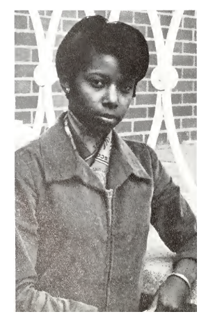 Venus Jones wearing a collared jacket is seated outdoors in front of a brick wall with a decorative metal railing