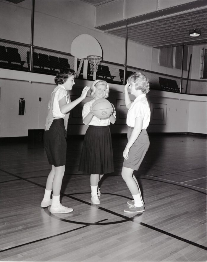 A young woman holding a basketball stands in between two other young women, preparing to toss the ball up for a jump ball.