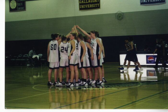 The women's basketball team stands in the middle of the court with their hands raised together during a huddle.