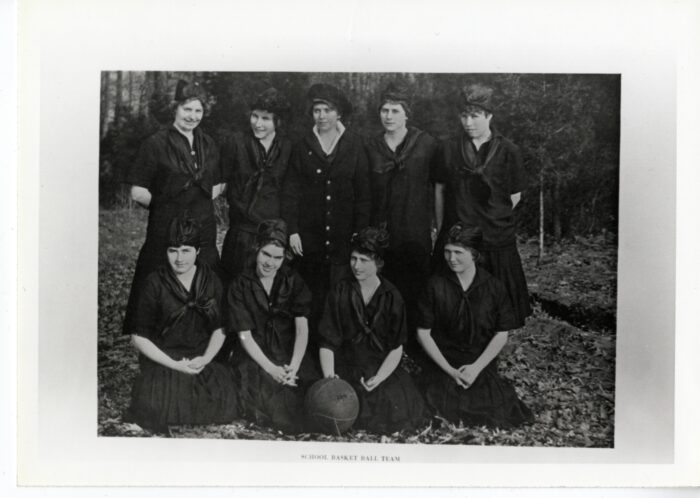 Nine girls from the basketball team pose - four sitting, five standing - while wearing long black dresses, neckerchiefs, and head scarves.