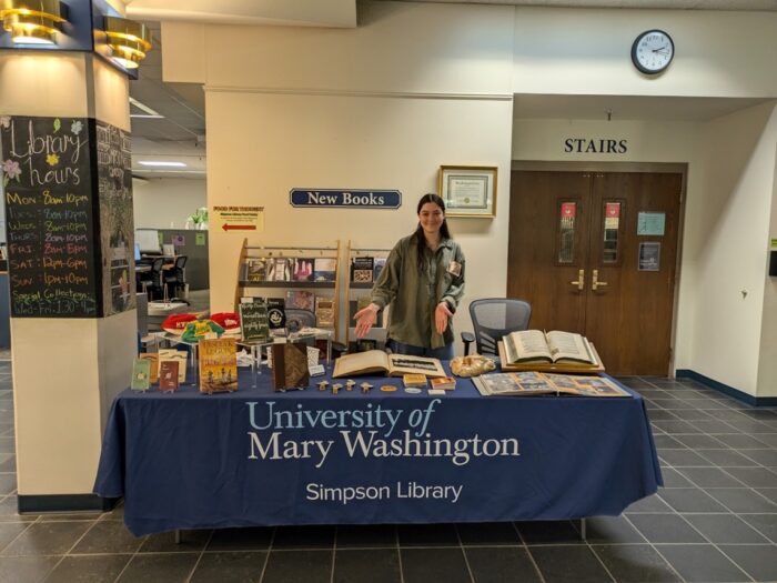 A person stands behind a table covered with a blue tablecloth. Books, small display items, and open volumes arranged across the surface.