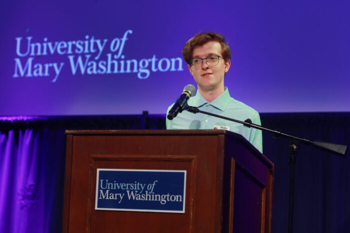 Person standing at a podium with a microphone, speaking on a stage with a projected “University of Mary Washington” logo in the background.