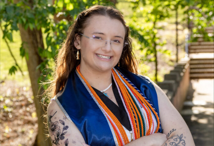 Person standing outdoors wearing graduation cords and a blue stole, with trees and stone steps in the background.