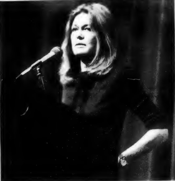 Gloria Steinem standing in front of microphone with a stage current behind her. Clipping from a newspaper