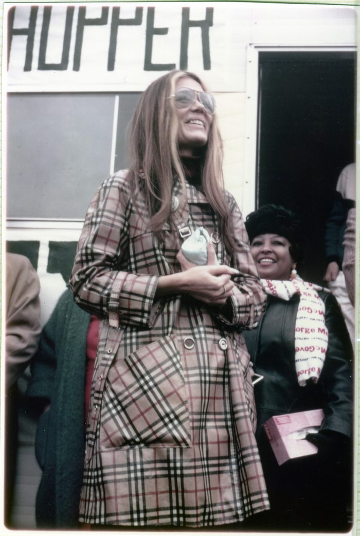 Gloria Steinem in an oversized aviator-style glasses and a long plaid coat, holding a small object. Several other people stand nearby. 