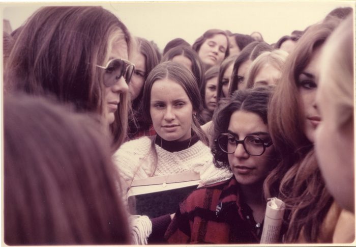 Gloria Steinem walking through crowd of college age students. Some are holding books and newspapers. Several people are blocking parts of the frame.