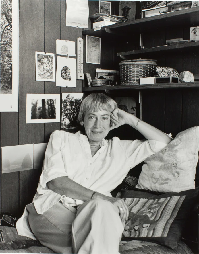 Photograph of a person seated on a cushioned bench in a wood-paneled room, surrounded by bookshelves, photographs, drawings, and decorative objects.